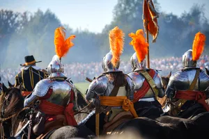 Honorable knights in armor with orange feathers and plumes riding on horseback, seen from the back with a view over the battlefield.