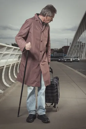 Man in trench coat with crutch on street.